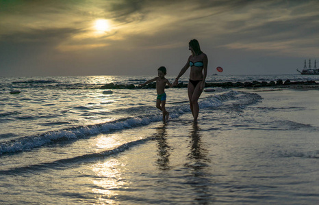年轻的母亲和微笑的小男孩儿子在日落的海滩上玩耍。 积极的人类情绪，感受快乐。 有趣可爱的孩子度假和享受夏天。 春夏假期。