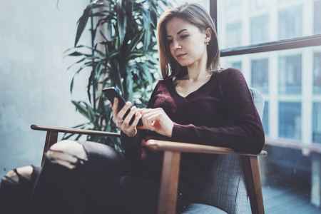 手持现代手机的金发青年女人。女孩指着手指在阳光明媚的 coworking 地方的空触摸移动屏幕。横向模糊背景