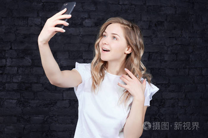 位年轻快乐的女人的肖像,手拿智能手机数码相机,在黑色砖墙背景下自拍