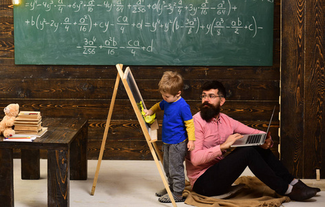 老师看着孩子擦黑板。 侧视男孩和男人坐在地上拿着笔记本电脑。 非正规教育概念。