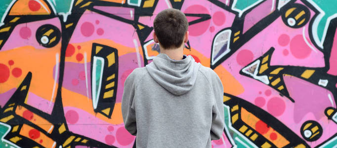 一位穿灰色连帽衫的年轻涂鸦艺术家在雨天的时候，用粉红色和绿色的涂鸦在墙上看着墙。街头艺术概念