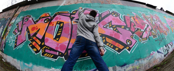 一个穿灰色连帽衫的年轻人在雨天在墙上画了粉红色和绿色的涂鸦。菲什耶枪