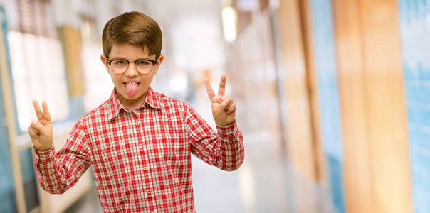 英俊的幼儿，绿眼睛，看着摄像机，用手指在学校走廊上做胜利标志