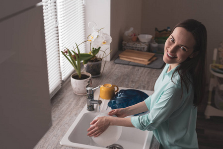 微笑的年轻女人在厨房里洗盘子前的一个窗口与鲜花在柔软的晨光中, 色调真实的场景角度从顶部和从侧面