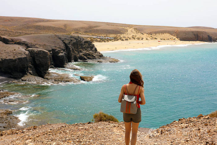 背着背包的年轻女人的背面欣赏兰萨罗特岛海岸的壮观景色,背景是海滩