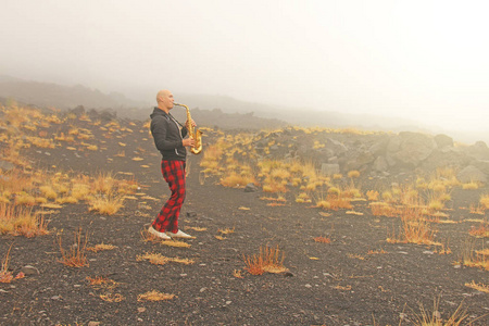 一个秃头男子在一个金色的女低音萨克斯在自然, 反对的背景下, 埃特纳火山火山, 在雾。浪漫的萨克斯爵士乐。这个人是个音乐家。意大