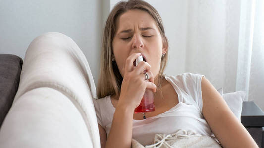 生病的年轻女子坐在沙发上, 使用喉咙喷雾的肖像