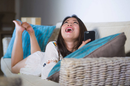 年轻美丽和快乐的亚洲华人妇女在她的20s 或30s 躺在客厅沙发沙发上使用互联网手机笑