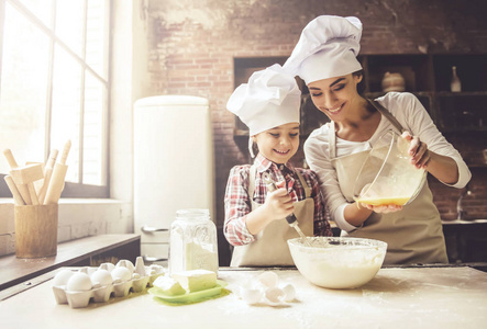 可爱的小女孩和她戴着厨师帽的漂亮妈妈在家烤东西时微笑着