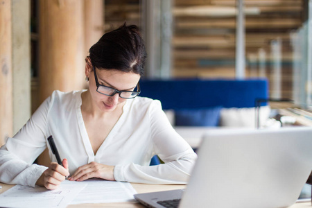商务女性，带着眼镜，在笔记本电脑后面工作，在舒适的咖啡馆氛围中，带着文件靠近窗户