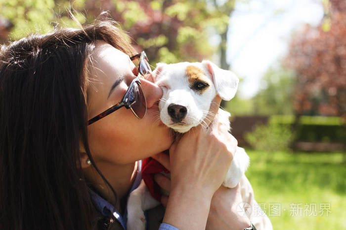 迷人的时髦的年轻女子在太阳镜亲吻杰克罗素猎犬小狗在公园绿色草坪和