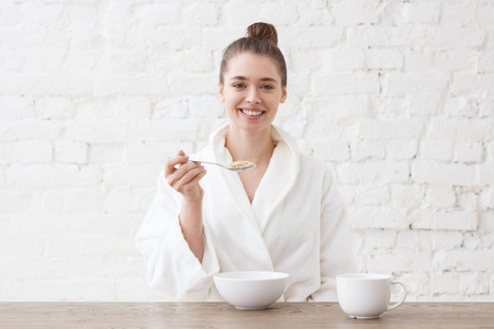 年轻的欧洲白种女人的室内图片坐在白色浴袍餐桌上吃谷物, 喝咖啡或茶早餐看着微笑的感觉积极和放松