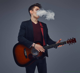 英俊的年轻音乐家与时髦的头发在优雅的衣服吹烟雾, 而演奏声学吉他