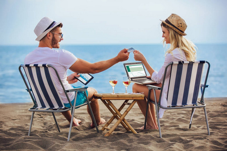 快乐的微笑夫妇上网，享受夏天在热带海滩使用笔记本电脑数字平板电脑和信用卡。