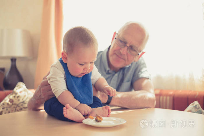 一位祖父坐在一张桌子旁，正在帮助他的小孙子学习如何吃固体食物