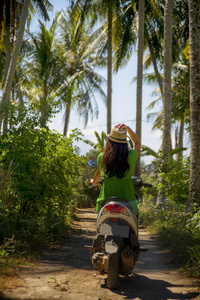 年轻快乐的旅游妇女与帽子骑摩托车摩托车在热带天堂丛林与蓝天和棕榈树探索旅行目的地