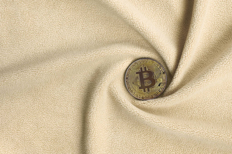黄金比特币躺在一条毯子上，由柔软蓬松的浅橙色羊毛织物制成，有大量的浮雕褶皱。 折叠的形状就像一个来自显卡冷却器的风扇