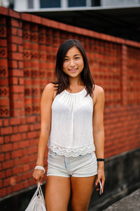 一位年轻迷人的新加坡华人亚洲妇女的肖像，她拿着电话站在一条靠砖墙的街道上。 她微笑着，身材娇小，运动，晒黑。