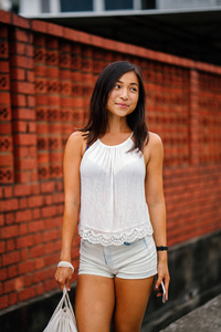 一位年轻迷人的新加坡华人亚洲妇女的肖像，她拿着电话站在一条靠砖墙的街道上。 她微笑着，身材娇小，运动，晒黑。