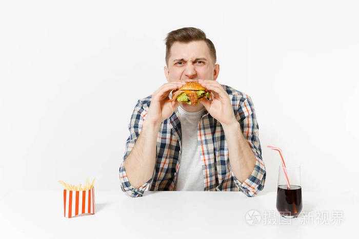 有趣的饥饿年轻人吃汉堡,坐在桌子上,薯条可乐在玻璃中,隔离在白色