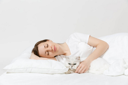 平静的年轻女人躺在床上，白色背景上有白色可爱的波斯银色下巴猫床单枕头毯。 美女花时间在房间里。 休息放松好心情的概念。 复制空间