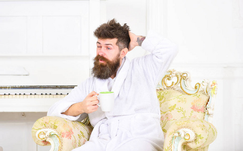 胡子和胡须的男人喜欢早上坐在老式的豪华扶手椅上。男子昏昏欲睡的浴衣喝咖啡在豪华酒店在早上, 白色的背景。上午咖啡概念