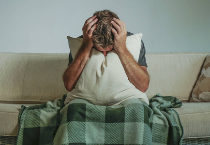 年轻的悲伤和绝望的男人坐在客厅沙发上,用手和枕头捂着脸,哭泣,痛苦