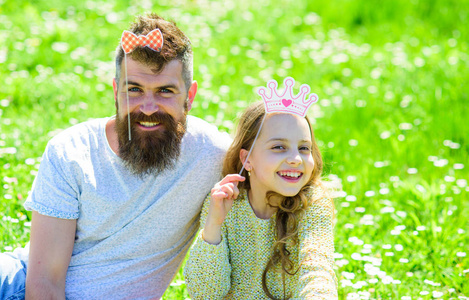 爸爸和女儿坐在草地上的草地, 绿色的背景。儿童和父亲摆着皇冠和弓照片展位属性。父亲的概念。家庭消费休闲户外, 玩少女游戏