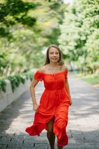 一位年轻迷人的高加索女人在一天的户外公园里沿着一条小径散步跑步的肖像。 她穿着一件漂亮的轻盈舒适的橙色连衣裙，笑得很开心。