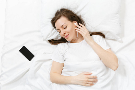 幸福的年轻女人躺在床上，用白色的床单枕毯，用手机听耳机里的音乐。 平静美丽的女性花时间在房间里。 放松，放松，放松心情