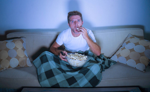生活方式肖像年轻迷人，放松的男人在电视上看电影，吃爆米花，深夜坐在客厅沙发沙发上，享受着完全集中和感兴趣的生活方式