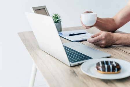 男子端着一杯咖啡在桌上，手提电脑课本相框盆栽和盘子上的甜甜圈的局部视图