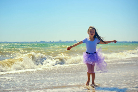 一个穿着紫色裙子的小女孩在阳光明媚的日子里在汹涌的海浪的背景下在海滩上跳舞。 肖像。 图像的水平方向。
