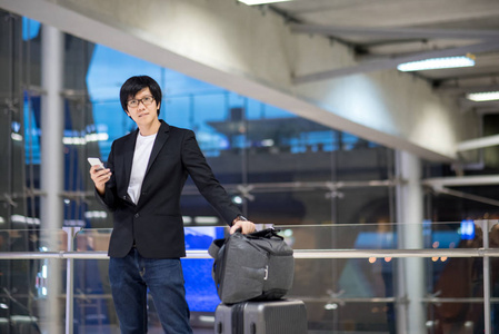 年轻的亚洲商人带着他的手提箱行李，使用智能手机，在国际机场候机楼等待航空航班，商务旅行和网上登记概念