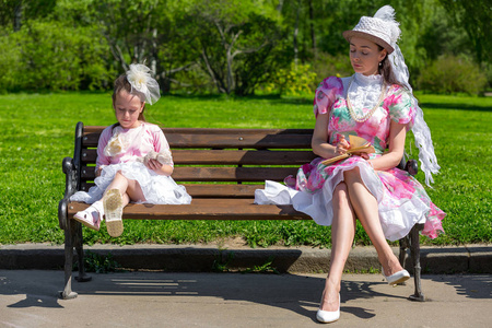 家庭在绿色公园的木凳上阅读旧书，穿着漂亮的复古连衣裙和优雅的帽子阅读书籍，这是关系和价值观的概念。 母亲和女儿在夏天的公园里。