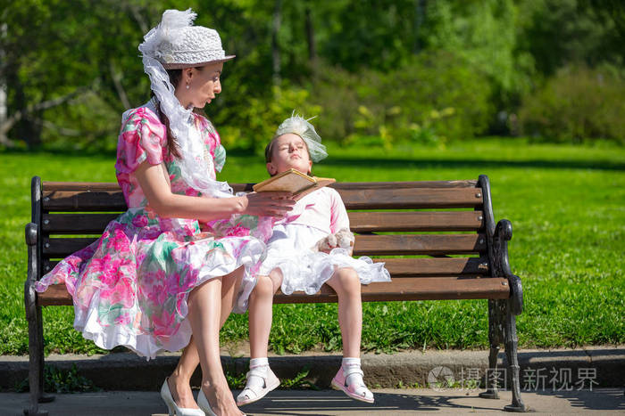 家庭在绿色公园的木凳上阅读旧书，穿着漂亮的复古连衣裙和优雅的帽子阅读书籍，这是关系和价值观的概念。 母亲和女儿在夏天的公园里。