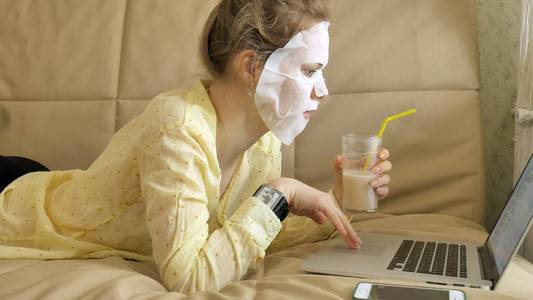 年轻妇女做面膜面具与清洁面具, 工作在笔记本电脑在家里