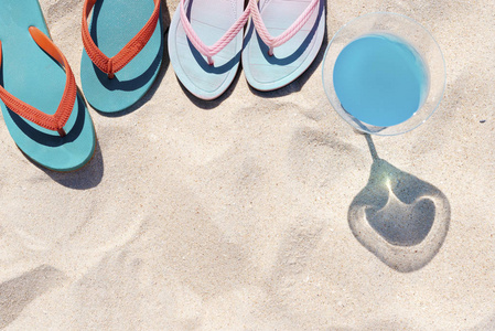 旅行和暑期派对。 五颜六色的拖鞋在海滩上，蓝色的饮料在白沙上。 暑假快乐。 图片用于添加文本消息。 设计艺术作品的背景。
