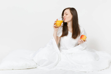 年轻的黑发女人坐在床上，白色的床单枕头包裹在毯子上，孤立在白色的背景上。 美女早餐吃苹果新鲜橙汁。 良好的情绪观念