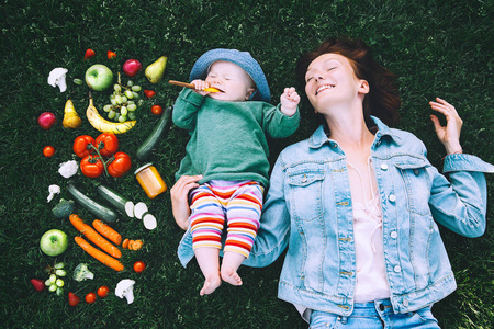 快乐的妈妈和小宝宝躺在绿草上，有不同的新鲜水果和蔬菜。 健康儿童的背景和父母营养的顶部视图。 家庭食物概念彩色照片