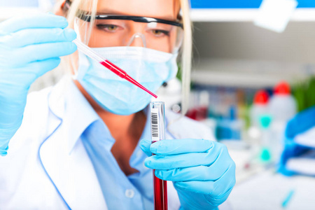 年轻有魅力的女科学家戴着防护眼镜和手套，在科学化学实验室里用长玻璃吸管把一种红色液体物质滴到试管里