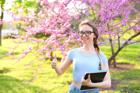 欢快的女学生在盛开的公园里竖起大拇指和保存文件