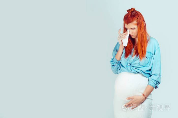 怀孕期间的情绪。年轻的随便女人摸着肚子里温柔的婴儿，用纸纸巾擦拭眼泪，俯视浅蓝色。混合种族模式，拉丁西班牙裔爱尔兰妇女