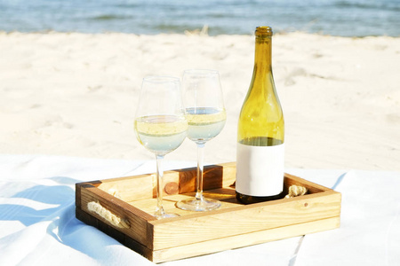 浪漫的晚餐构图，昂贵的一瓶白葡萄酒空白标签和两杯木托盘在白色沙滩海洋。 海景。 关闭背景复制空间。