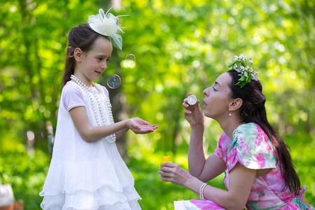 妈妈和女儿一起玩，在一个阳光明媚的绿色公园里推出肥皂泡，充满了乐趣和欢乐。 白色复古连衣裙与帽子和服装珠宝。 夏天公园里的家人。