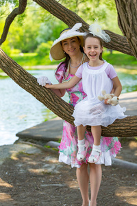 女儿坐在树枝上，笑着穿着白色的浅色连衣裙。 妈妈站在一顶漂亮的帽子旁边，戴着手套拥抱。 母爱的肖像。 夏天公园里的家人。