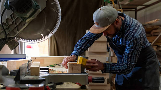 有经验的木工在工作服和小木匠老板在木工车间工作，用砂纸打磨桌子上的工作台上的鸟舍，是一个锤子圆锯和许多工具