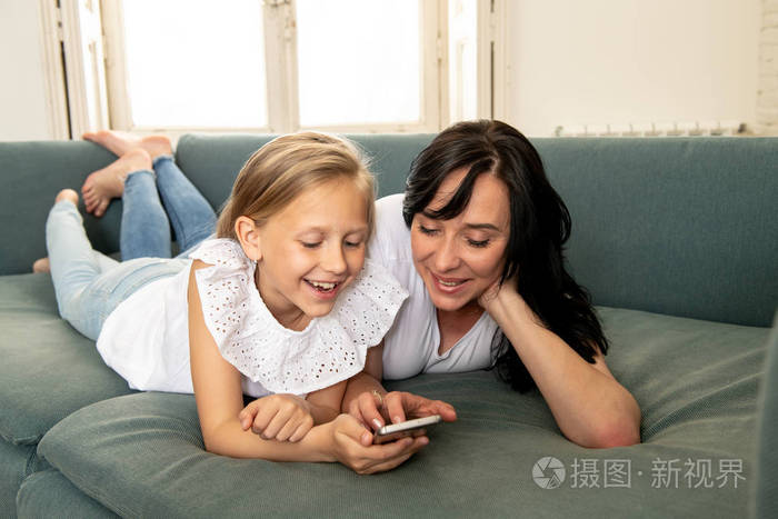 漂亮的金发可爱的小女孩和妈妈一起玩，微笑着，在家里的沙发上用智能手机进行亲密接触。 幸福家庭数字技术和生活方式理念。