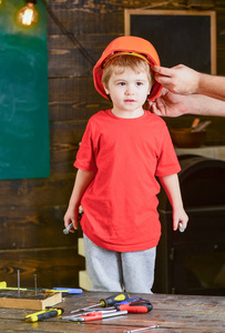 幼儿在保护硬帽子, 头盔在家里的车间。孩子可爱和可爱的立场, 而男性手把硬帽子头上。小心地保护孩子的头盔。保护和安全概念