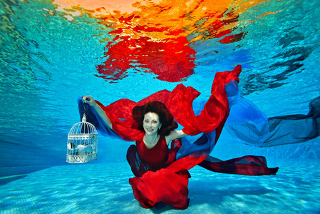 一个年轻的女孩在池底水下游泳,玩红色和蓝色的布料,手里拿着一个白色